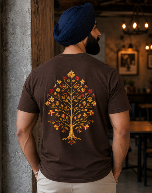 A man wearing a dark brown t-shirt with a large golden tree of life graphic on the back, paired with a navy blue turban and beige pants, standing in a rustic indoor setting.