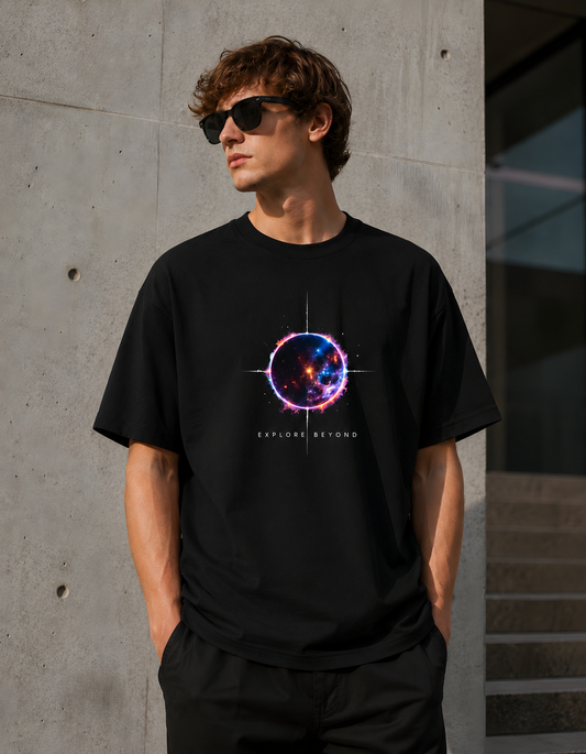A man wearing a black graphic t-shirt with a colorful cosmic ring design and the text 'EXPLORER BEYOND' on the front, standing against a concrete wall.