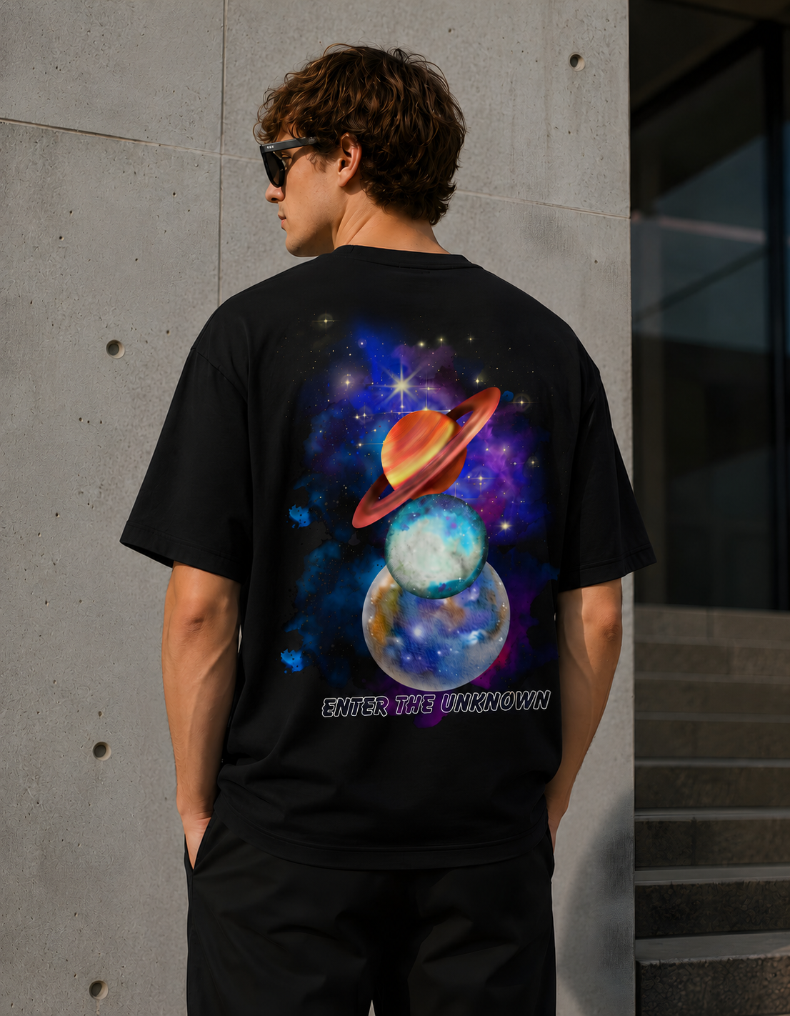 A man wearing a black t-shirt with a colorful cosmic graphic of planets and stars, standing in front of a concrete wall.