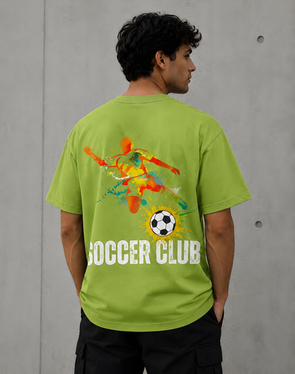 A man wearing a lime green t-shirt from the back, featuring a colorful watercolor soccer graphic with the text 'SOCCER CLUB' in white. The background is a textured gray concrete wall.