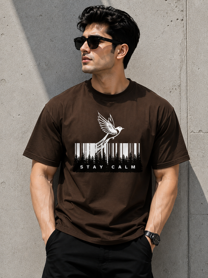A man wearing a dark brown crewneck t-shirt with a white graphic of a bird flying over a barcode and the text 'STAY CALM', posed against a textured concrete wall.