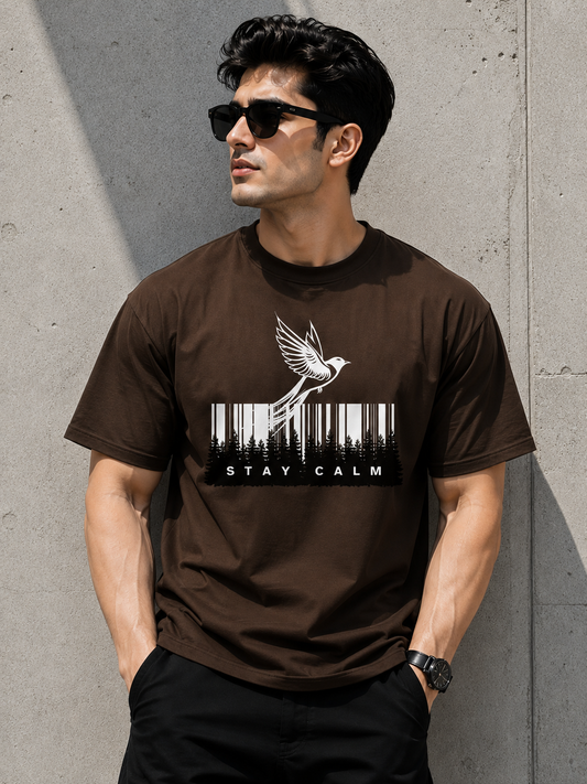 A man wearing a dark brown crewneck t-shirt with a white graphic of a bird flying over a barcode and the text 'STAY CALM', posed against a textured concrete wall.