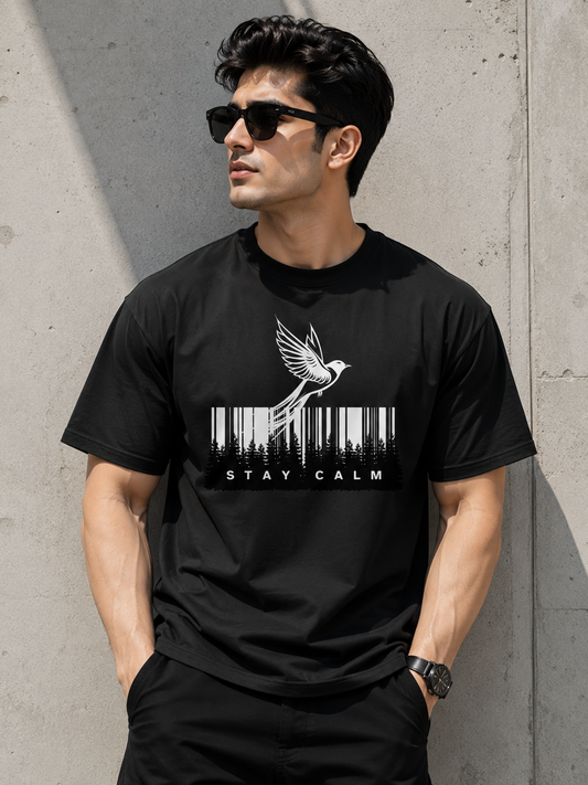 A man wearing a black crewneck t-shirt with a white graphic of a bird flying over a barcode and the text 'STAY CALM', posed against a concrete wall.
