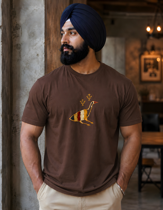 A man wearing a dark brown crewneck t-shirt with a colorful graphic of a bird on a surfboard, paired with beige pants and a navy blue turban, standing in a rustic indoor setting.