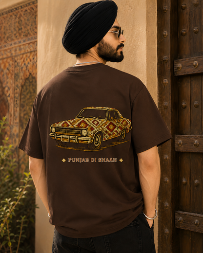 A man wearing a black turban and glasses, viewed from behind, wearing a brown t-shirt with a graphic of a car decorated with a Punjabi-style geometric pattern and the text 'PUNJAB DI BHAAG'.