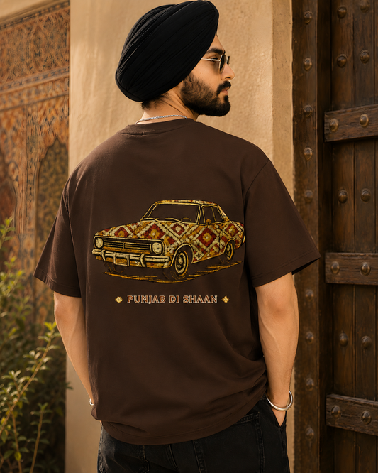 A man wearing a black turban and glasses, viewed from behind, wearing a brown t-shirt with a graphic of a car decorated with a Punjabi-style geometric pattern and the text 'PUNJAB DI BHAAG'.