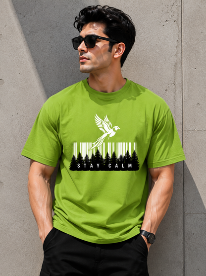 A man wearing a bright lime green t-shirt with a black and white graphic of a bird flying over a forest and the words 'STAY CALM', standing against a concrete wall.