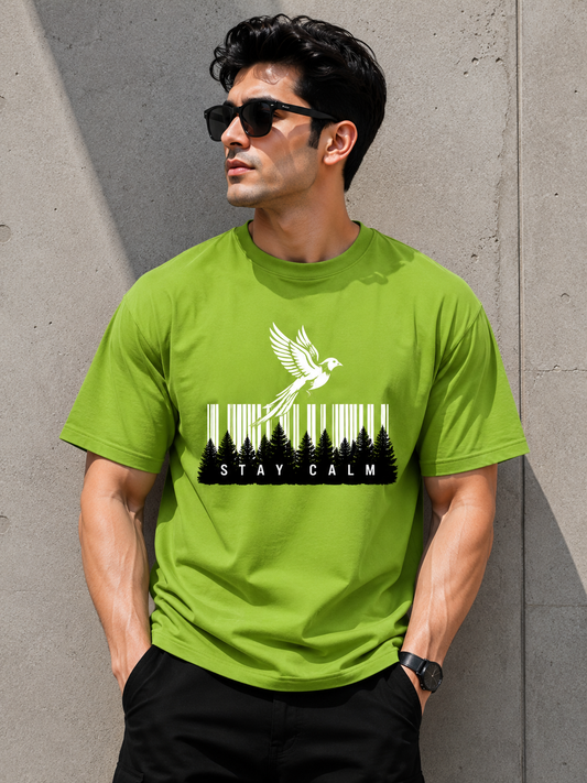 A man wearing a bright lime green t-shirt with a black and white graphic of a bird flying over a forest and the words 'STAY CALM', standing against a concrete wall.