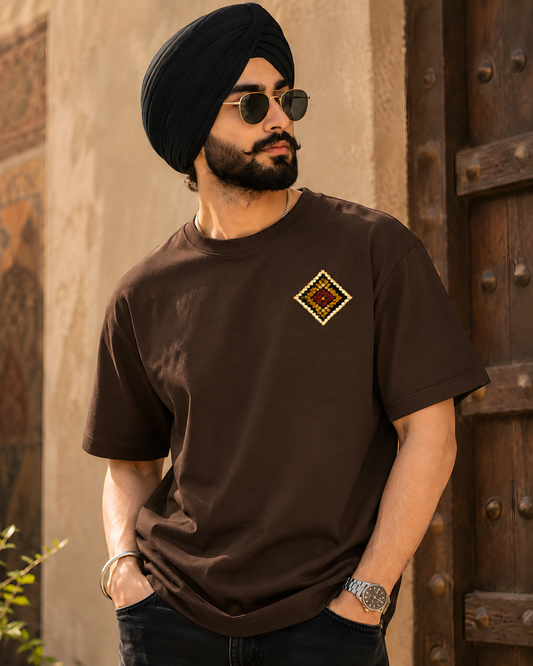 A man wearing a black turban, round sunglasses, and a dark brown short-sleeve t-shirt with a small geometric diamond embroidery on the left chest, standing in front of a rustic wooden door and beige wall.